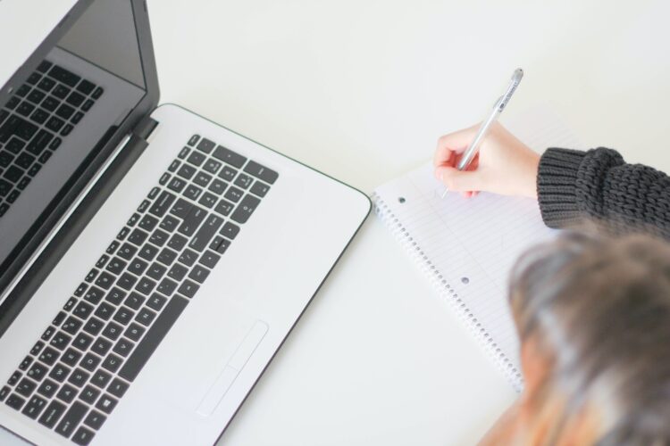 silver laptop and person taking notes on a sheet of paper next to it